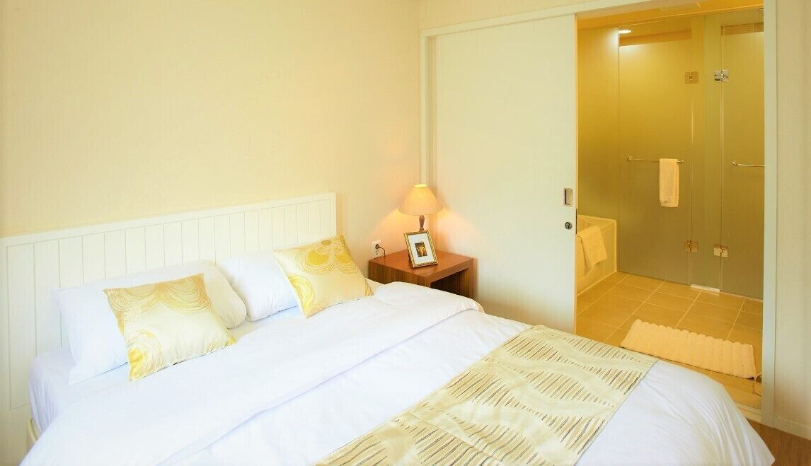 Ein Hotelzimmer mit einem Bett, einem Nachttisch mit Lampe, einem Badezimmer mit Duschkabine und einer Tür zu einem weiteren Raum.
