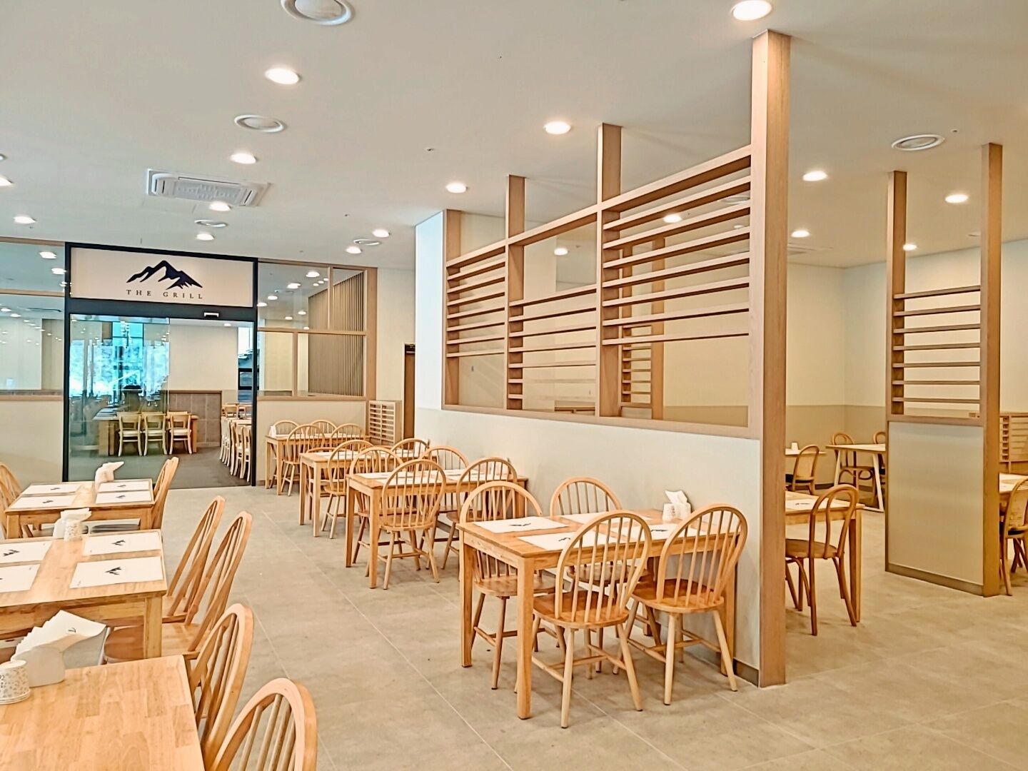 A restaurant interior with wooden tables and chairs, a large window with blinds, and a sign reading "The Grill".