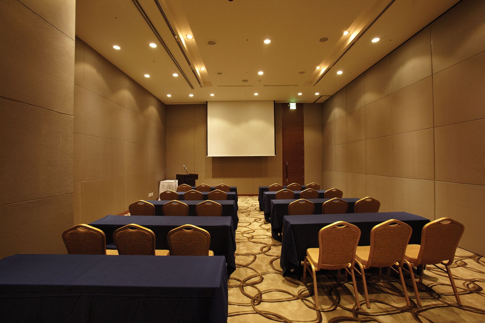 A conference room with rows of tables and chairs, a podium, and a projection screen.