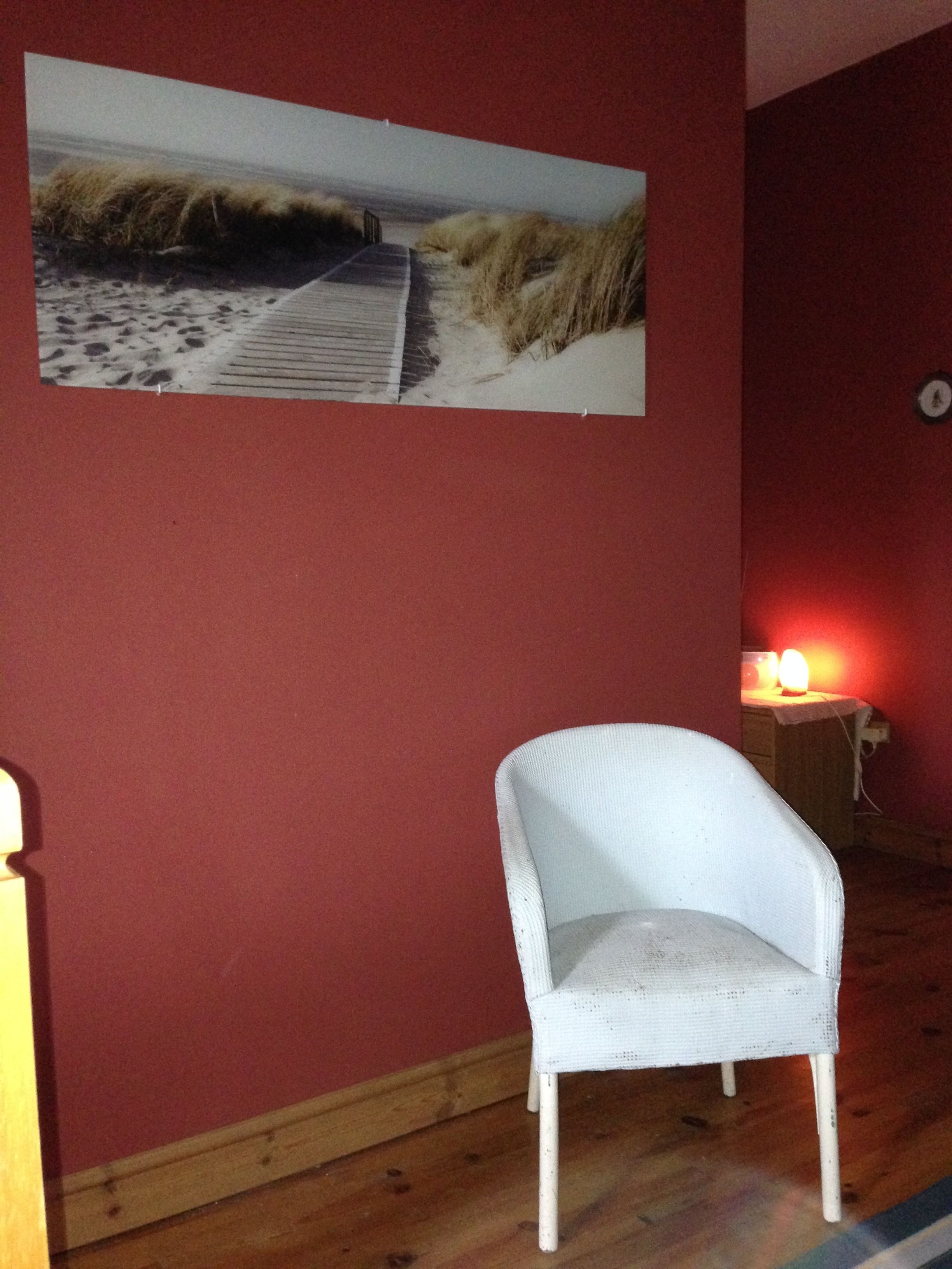 A room with a white chair, a red wall, and a painting of a beach scene.