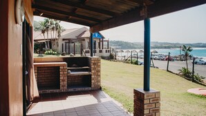 An outdoor patio with a brick barbecue area, a covered seating area, and a view of the ocean and beach. - Port Edward Holiday Resort (Port Edward)