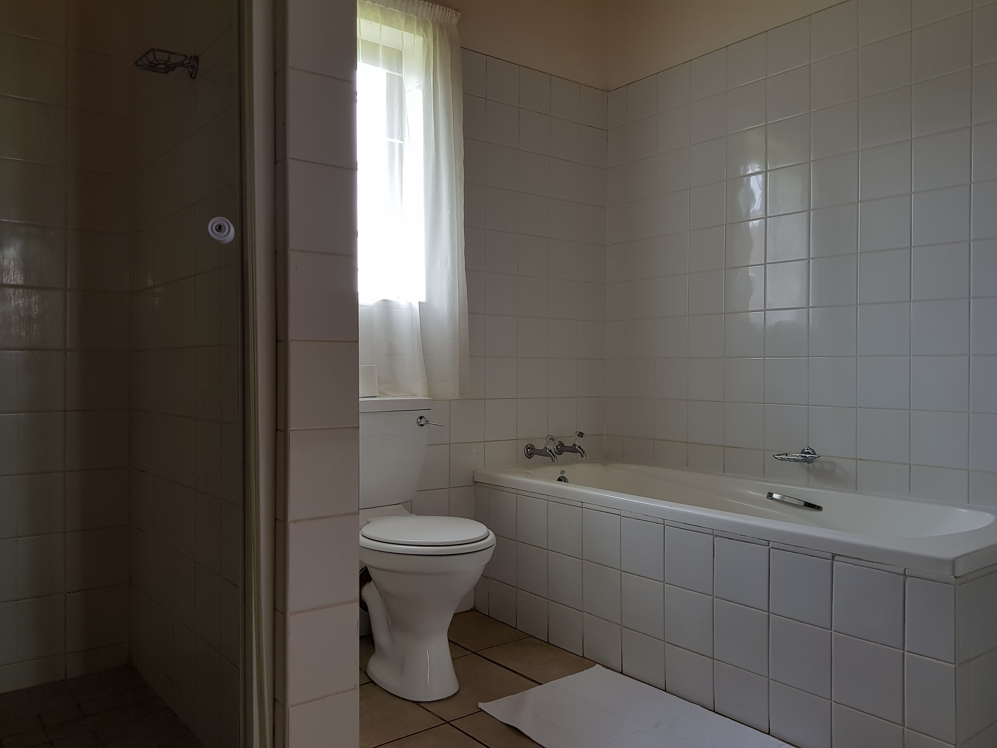 A bathroom with white tiles, a toilet, a bathtub, and a window with a curtain.