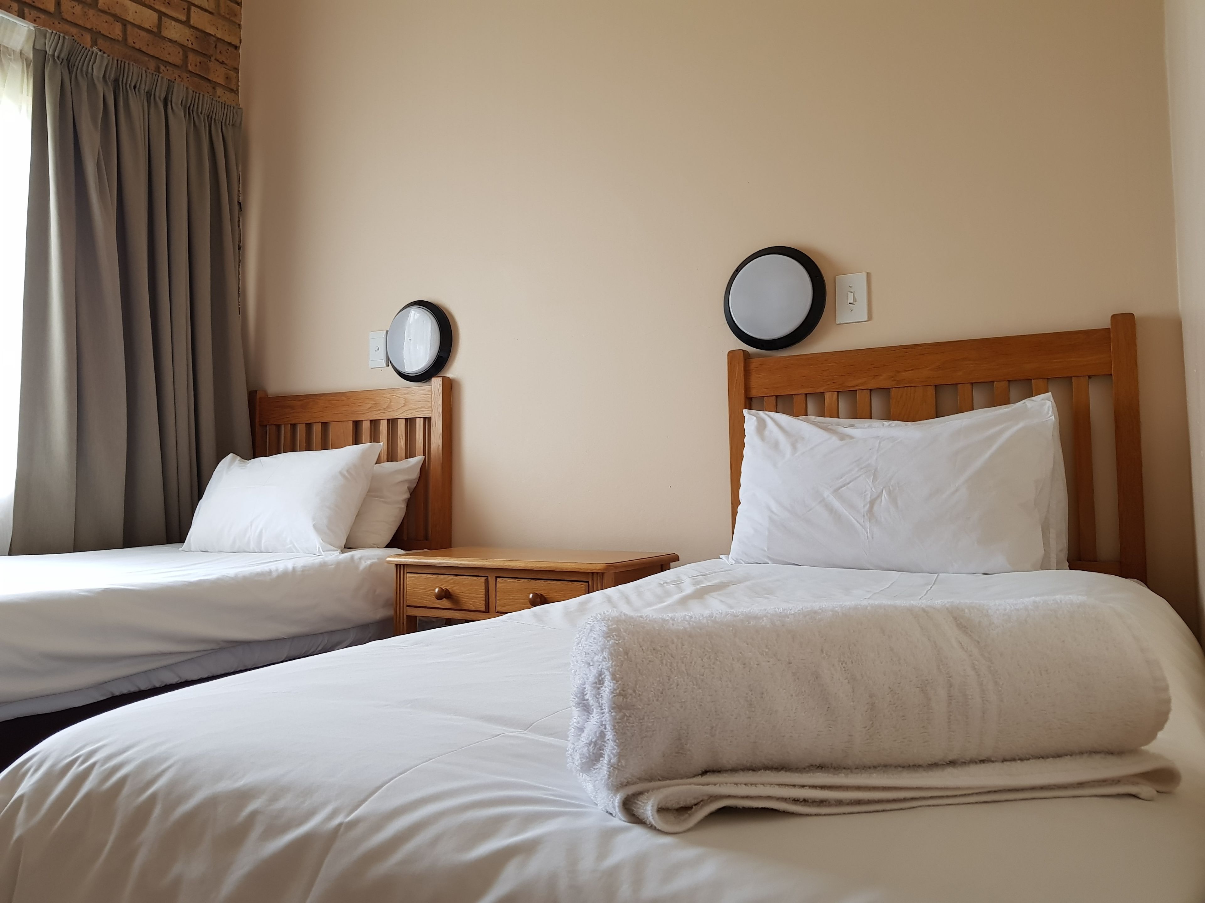 A hotel room with two single beds, wooden headboards, a nightstand with a lamp, and a wall clock.