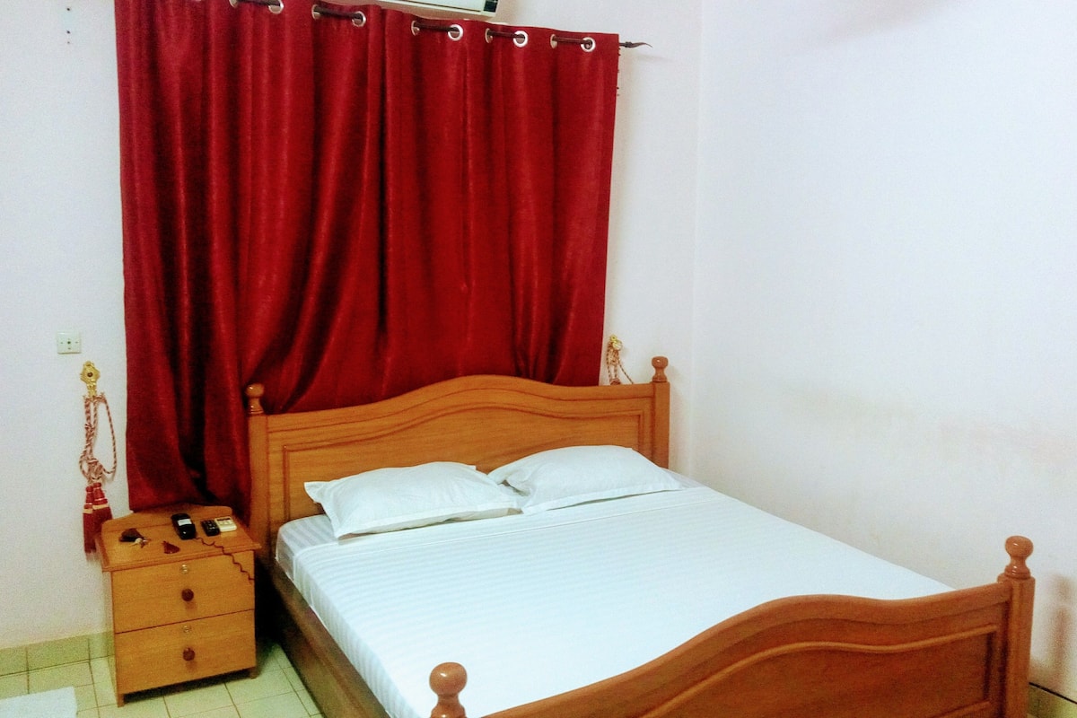 Una cama individual con cabecera de madera, una mesita de noche con lámpara y una cortina roja.