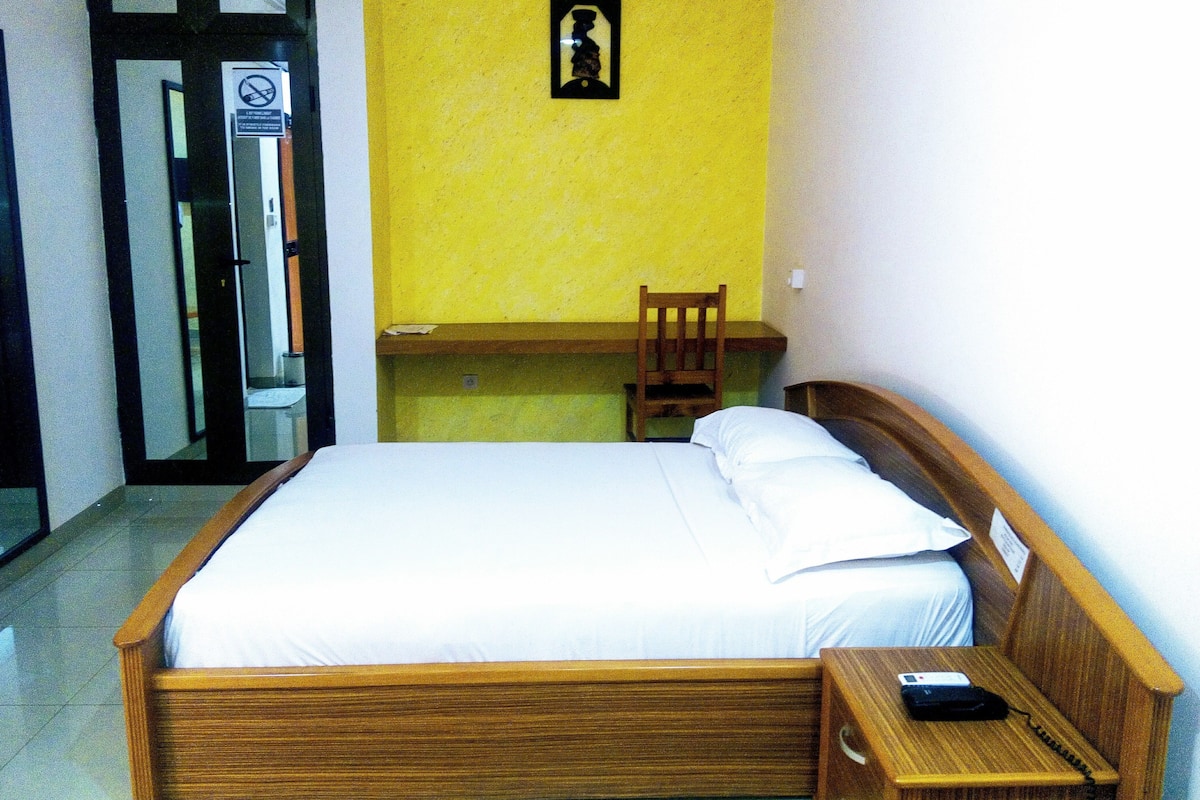 Una habitación con cama, mesita de noche de madera, una silla y una pequeña mesa.