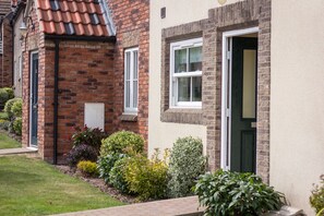A brick house with a green door, white windows, and a small garden with various shrubs.
