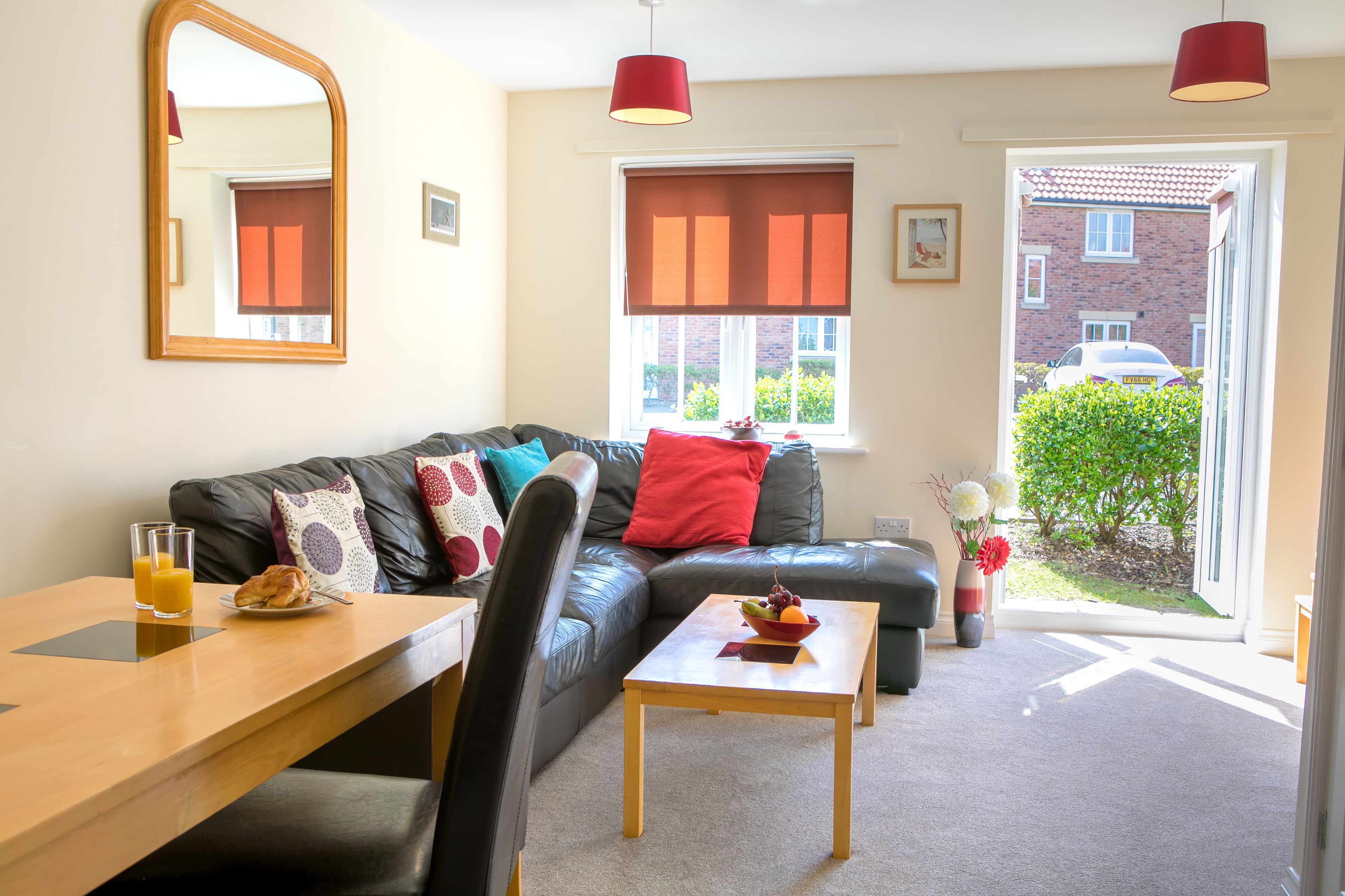 A modern living room with a leather sofa, a wooden coffee table, and a dining table with breakfast items. There is a large mirror on the wall, a window with blinds, and a view of a brick building outside.