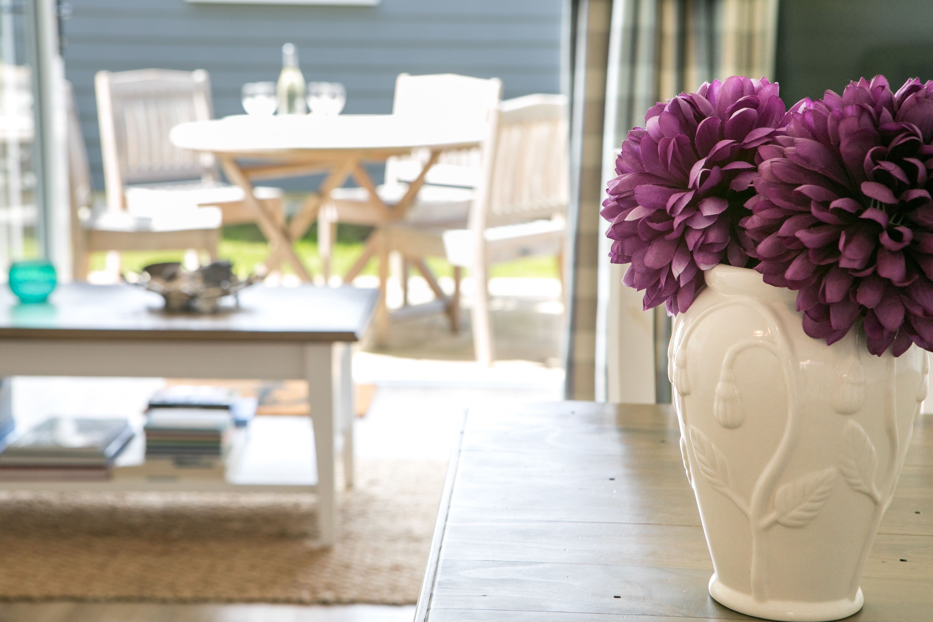 Un vase blanc orné de fleurs violettes, posé sur une table en bois dans une pièce dont on aperçoit, au fond, un coin repas.