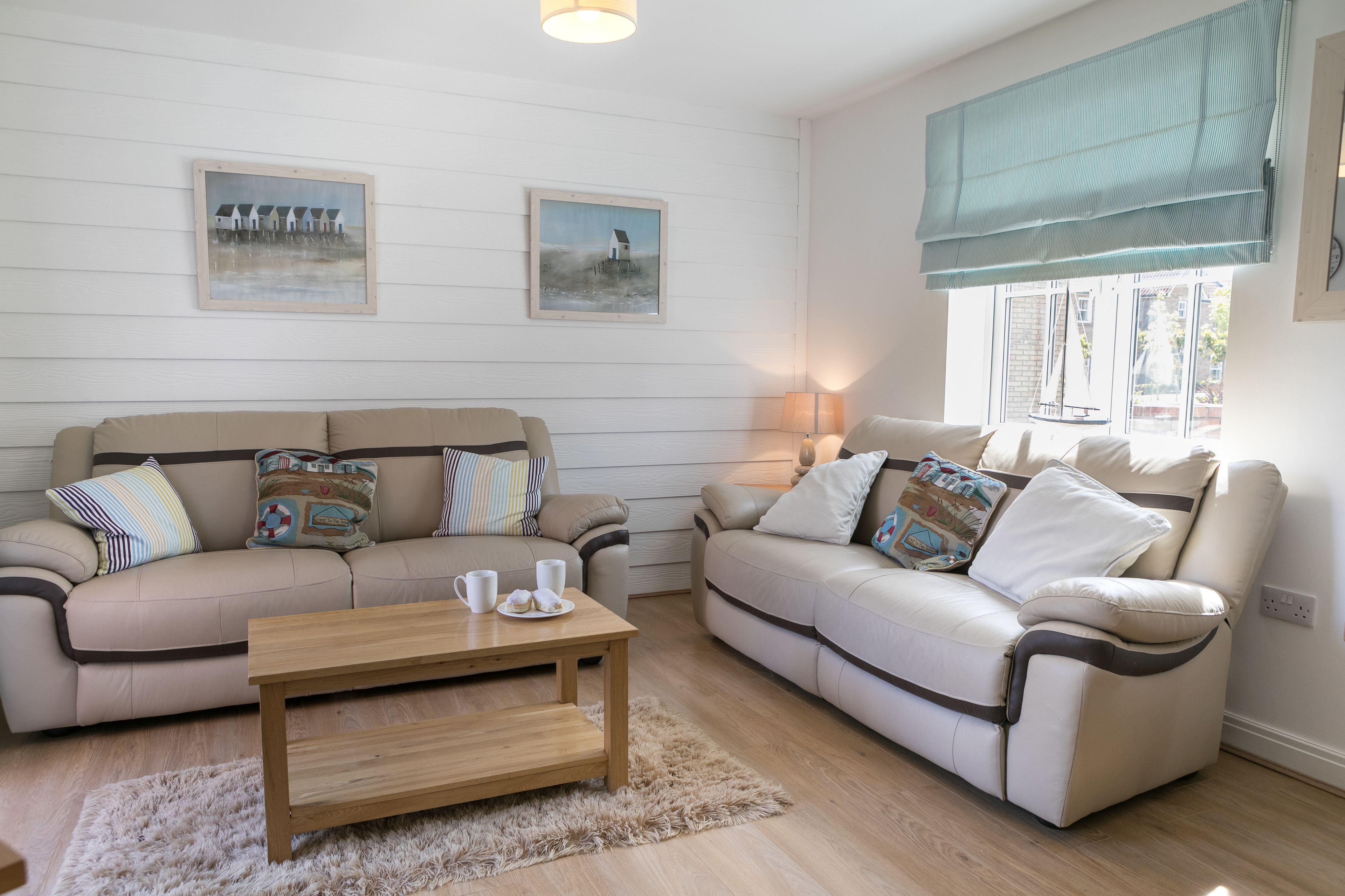 A living room with two beige sofas, a wooden coffee table, and a fluffy rug. There are two framed paintings on the wall and a window with a blue roller blind.