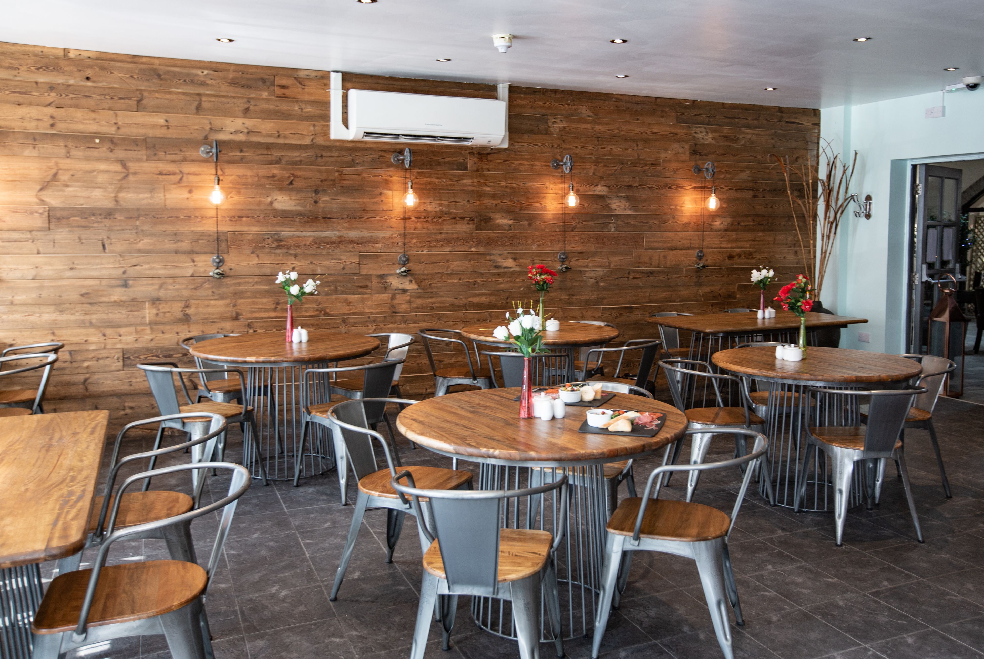 Un restaurante con mesas redondas y sillas de metal, paredes de madera y lámparas colgantes.