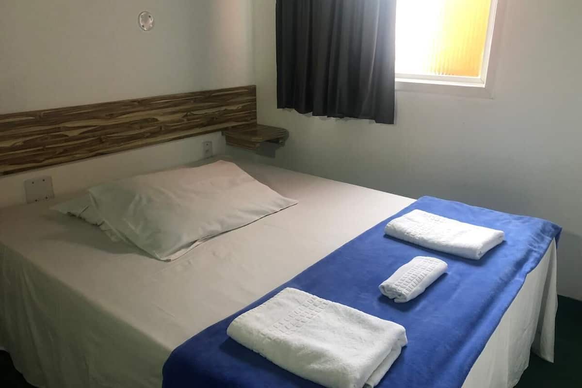 Una cama individual con cabecera de madera, una colcha azul, dos toallas blancas y una ventana con cortina.