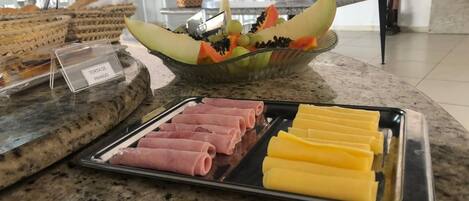 Una mesa de bufé con jamón en rodajas, queso y un bol de fruta con uvas, melón y piña.