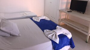 Habitación de hotel con dos camas, una con una manta azul y la otra con una blanca. Hay un televisor sobre un mueble de madera y un armario al fondo.