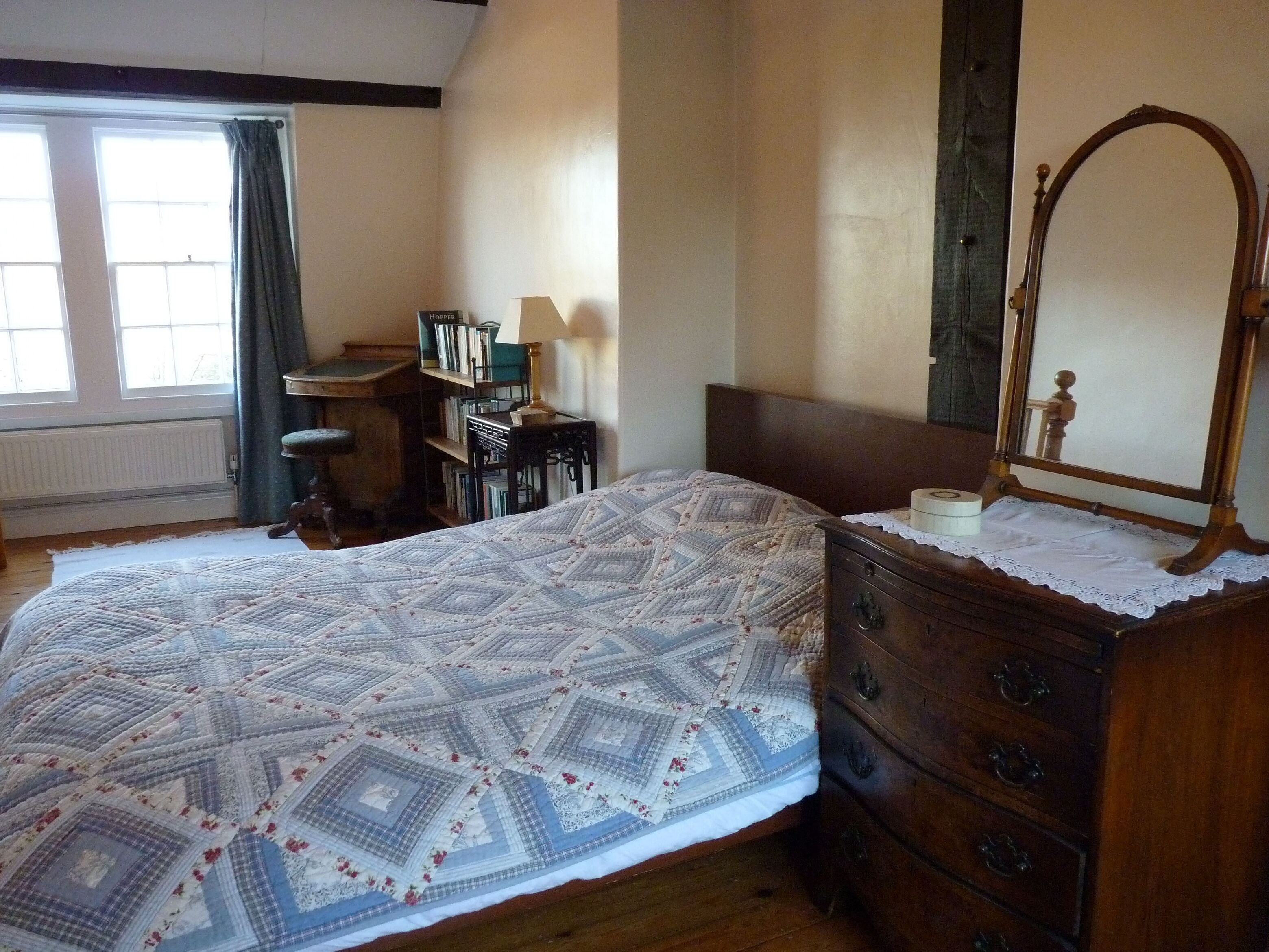 A bedroom with a large bed, a wooden dresser, a mirror, a desk with books, and a window with curtains.