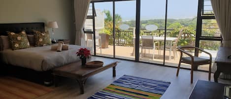 A bedroom with a large bed, a bedside table with a lamp, a wooden nightstand, a potted plant, and a view of a balcony with outdoor furniture.