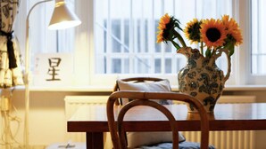 Una habitación con una silla de madera, una mesa, un florero con girasoles, una lámpara y una ventana con persianas.