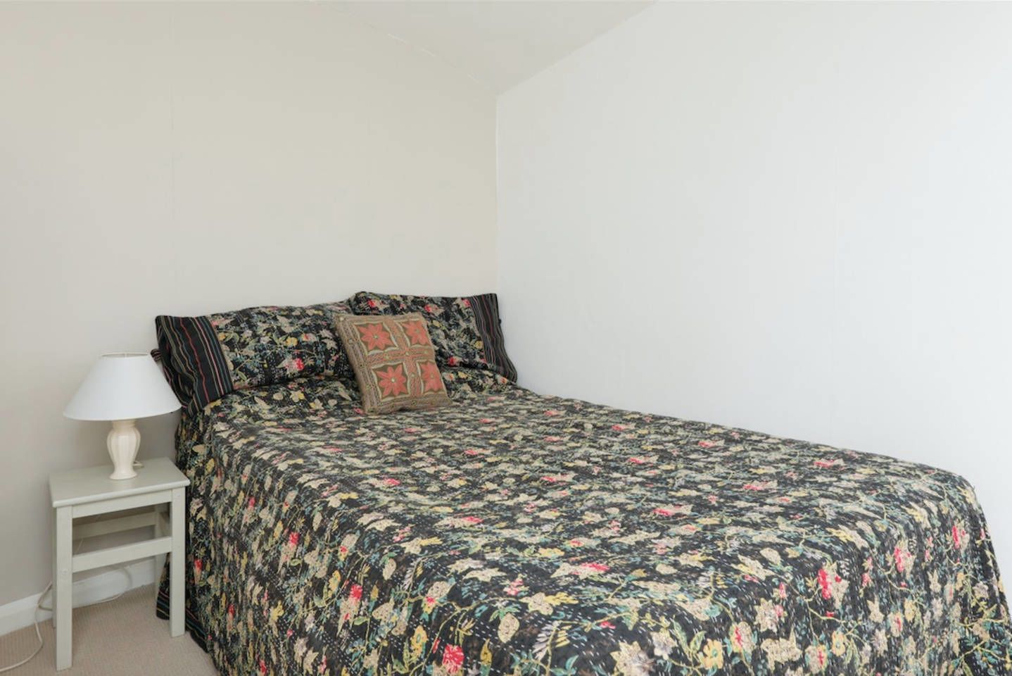 A bedroom with a floral bedspread, a white nightstand with a lamp, and a single bed with two pillows.