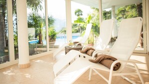 Un salón luminoso con sillas blancas y una mesita, con vistas a una piscina y abundante vegetación.