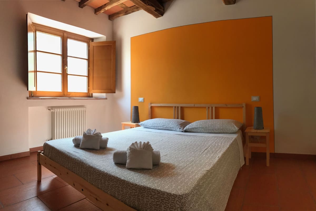 Een netjes opgemaakt bed met handdoeken, een raam met houten luiken en een gebogen oranje wand.