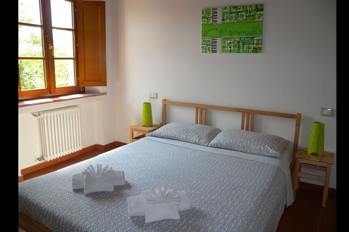 Een net opgemaakt bed met een bedsprei met patroon, twee nachtkastjes met lampen, een radiator en een raam met gordijnen.