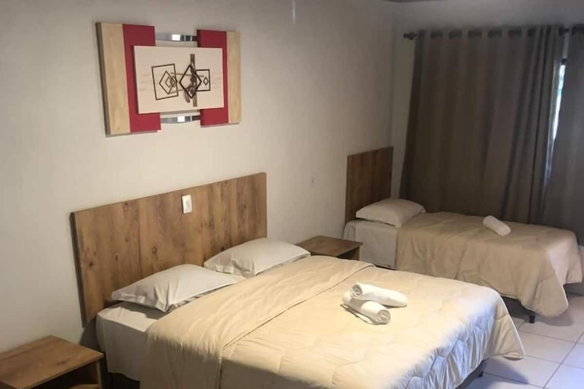 Een hotelkamer met een groot bed, twee kleinere bedden, een houten hoofdbord, een nachtkastje, een plank en een decoratie aan de muur.