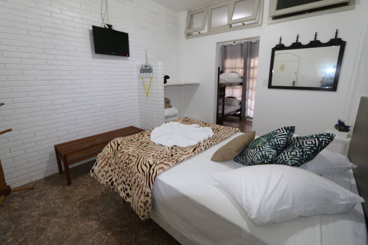 Een slaapkamer met een witte baksteenmuur, een houten bankje, een bed met beddengoed met een patroon, een spiegel en een aan de muur bevestigde televisie.
