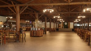 Een ruime eethoek binnen met houten tafels en stoelen, hoge plafonds met zichtbare balken en hanglampen.