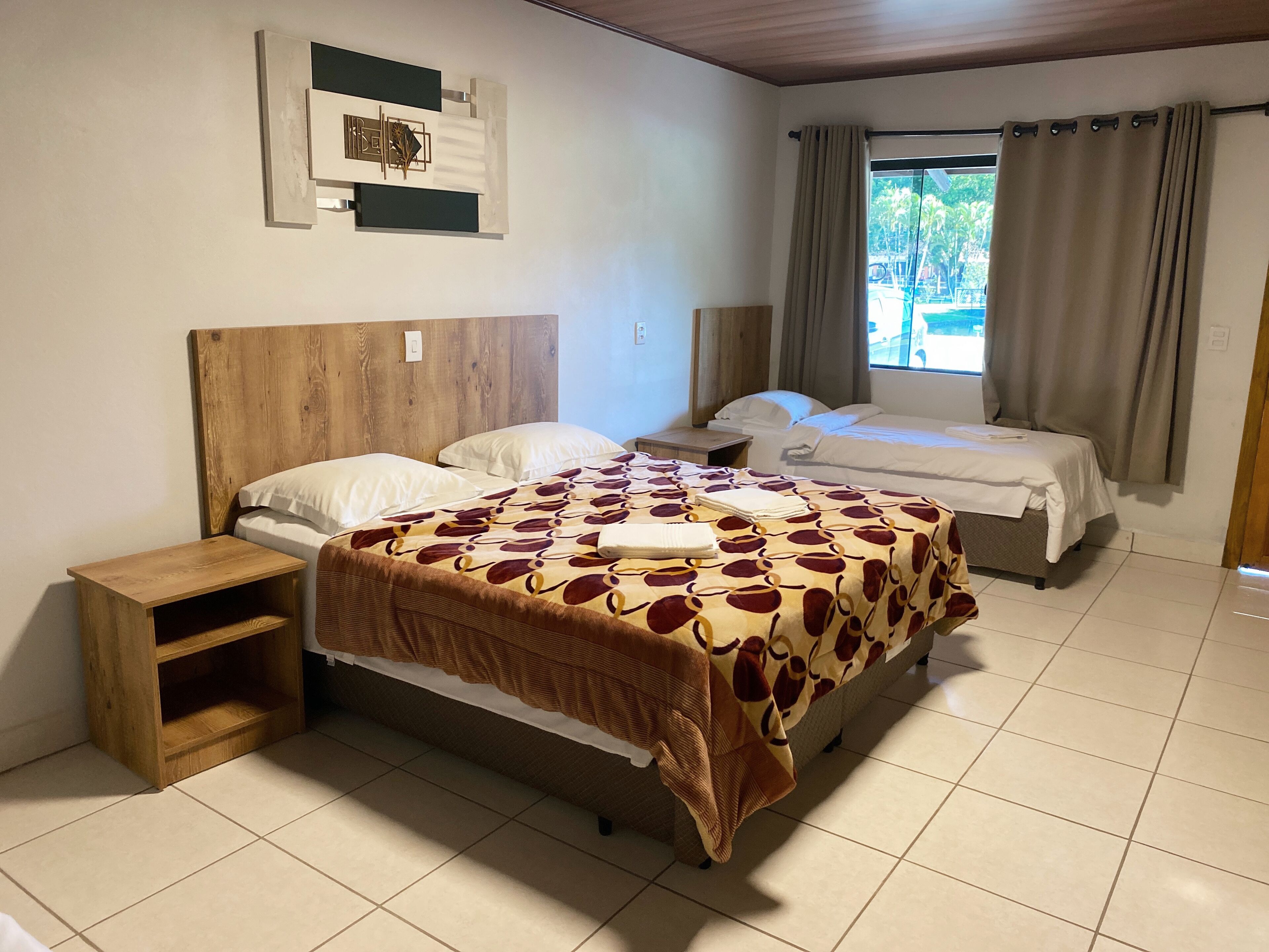 A hotel room with a large bed, two smaller beds, a wooden headboard, a bedside table, a shelf, and a window with curtains.