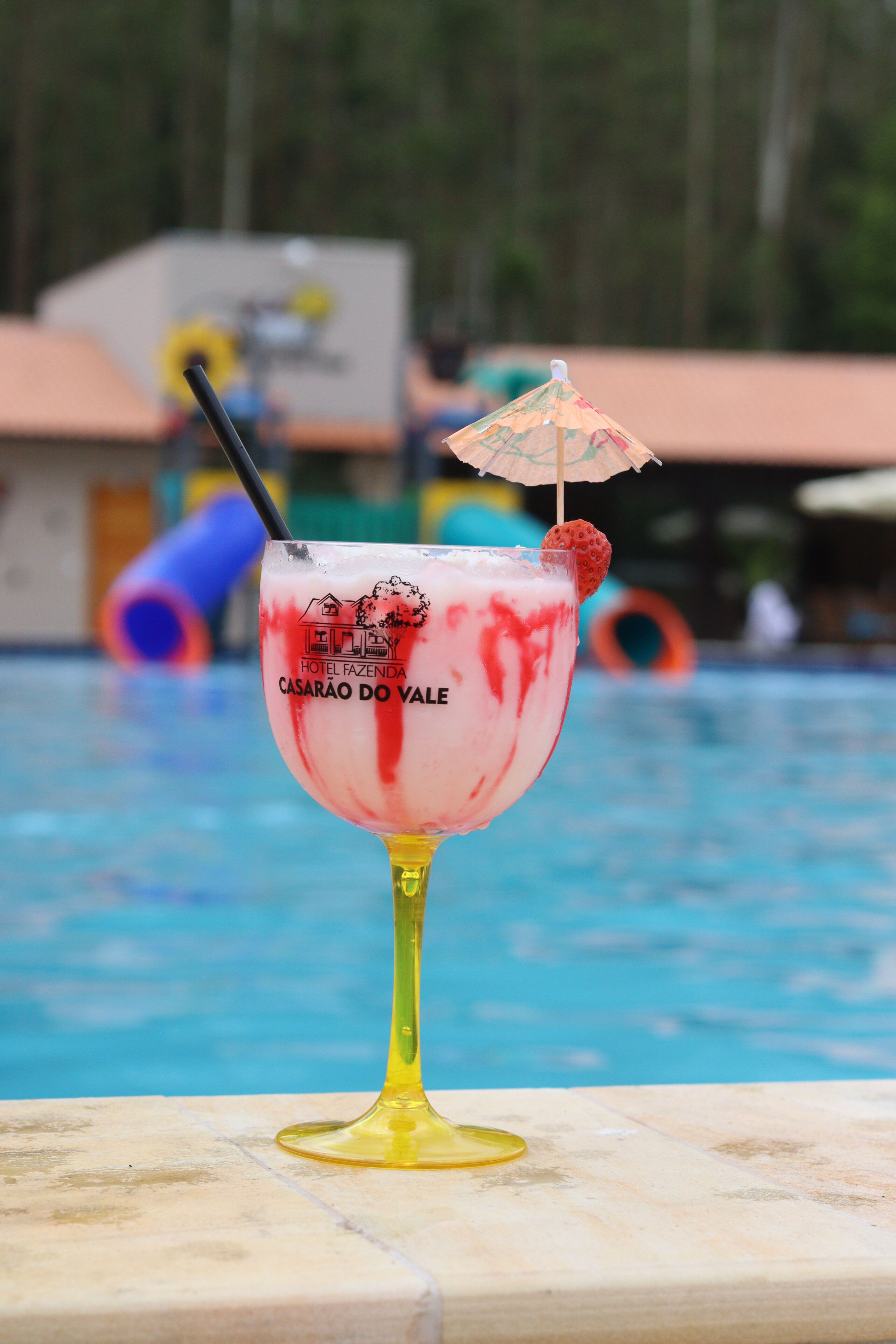 Un cocktail con una cannuccia e una fragola, con l'etichetta "CASARÃO DO VALE", davanti a una piscina, sullo sfondo un edificio e uno scivolo sfocati.