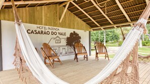 Een hangmat en houten stoelen onder een houten overkapping, met een muur waarop 'Hotel Fazenda Casarão do Vale' staat.