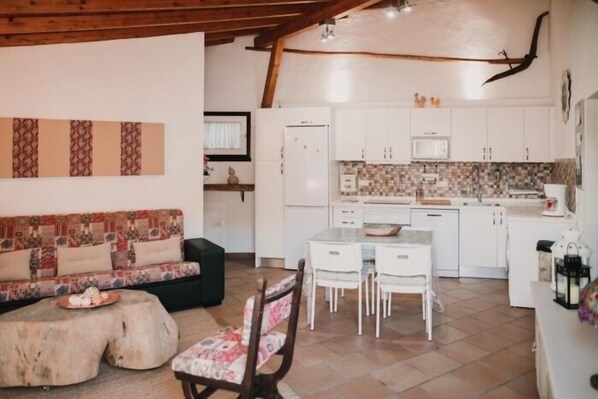 A cozy living area with a sofa, coffee table, and chairs, a kitchen with white cabinets and a tiled backsplash, and a wooden ceiling beam.
