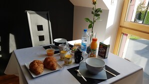 Een ontbijttafel met croissants, een vaas met een roos en diverse ontbijtartikelen, waaronder een fles water, sap en een pak Chocopic.