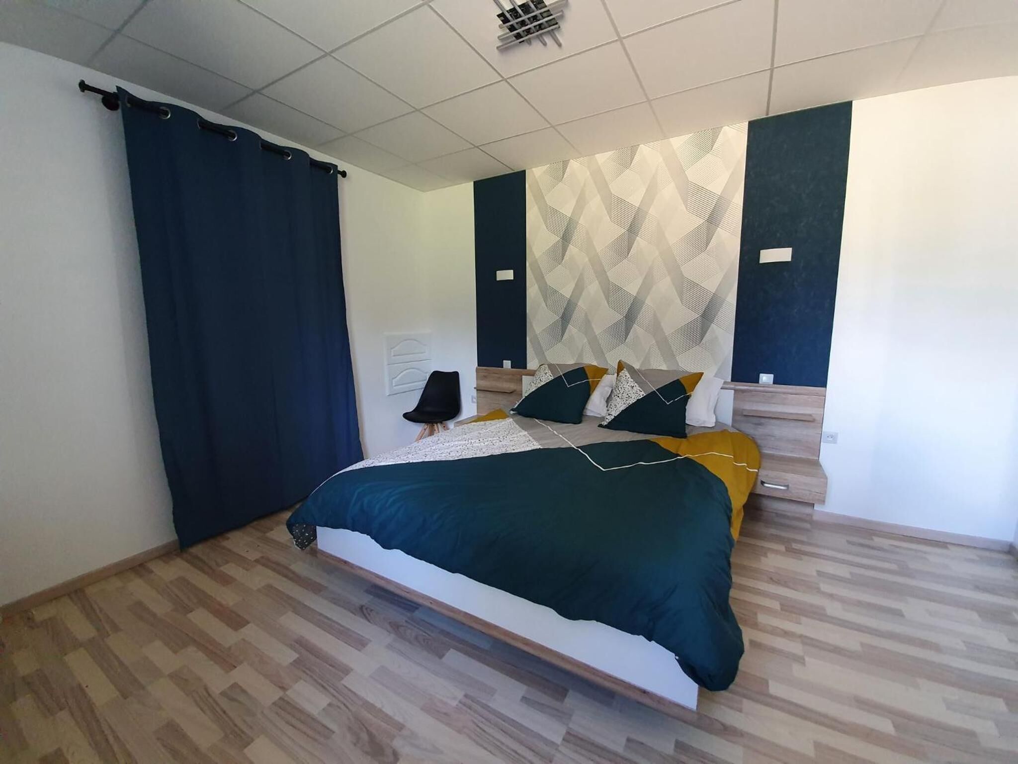 A modern bedroom with a bed, wooden flooring, a dark curtain, and a patterned wall.