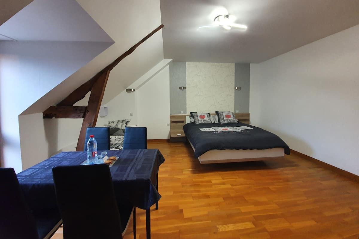 Quarto com cama, escrivaninha com cadeira, mesa com garrafa e copos, e ventilador de teto.