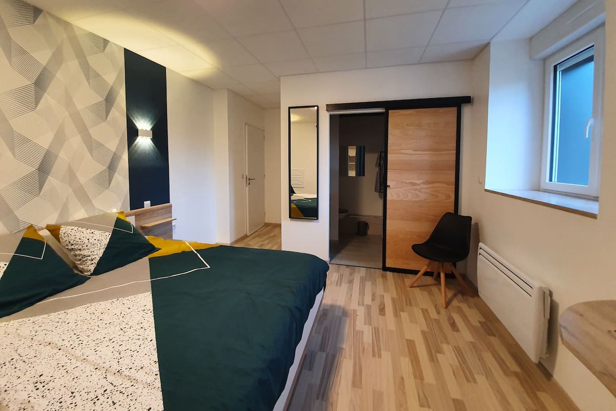 Quarto moderno com uma cama, uma cadeira, um guarda-roupa e um banheiro visível por uma porta aberta.