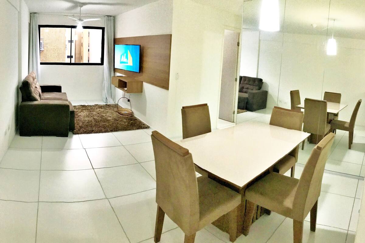A modern dining area with a rectangular table and six chairs, a sofa, and a television mounted on the wall.