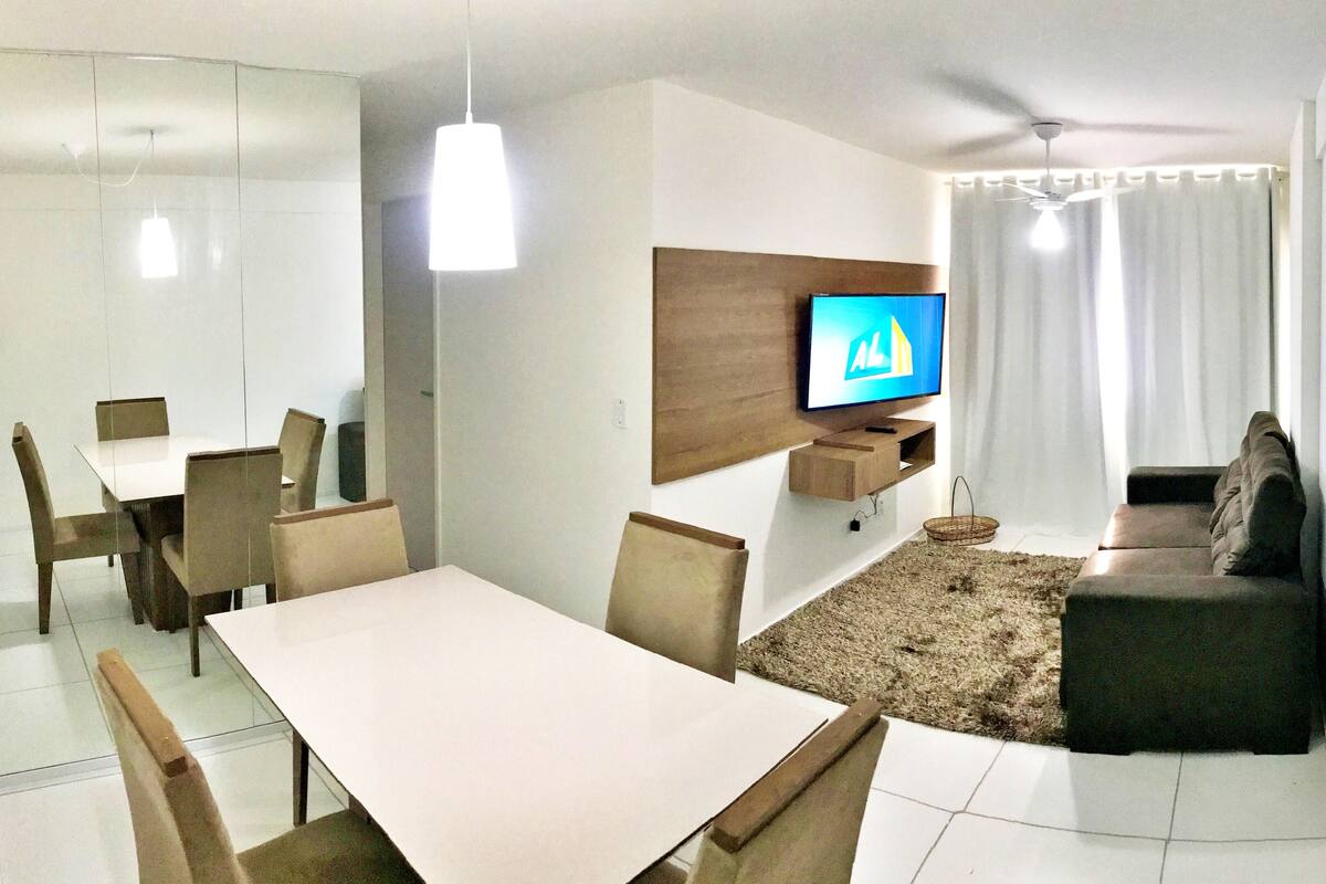 A modern living room with a flat-screen TV mounted on the wall, a dining area with a table and chairs, and a sofa.