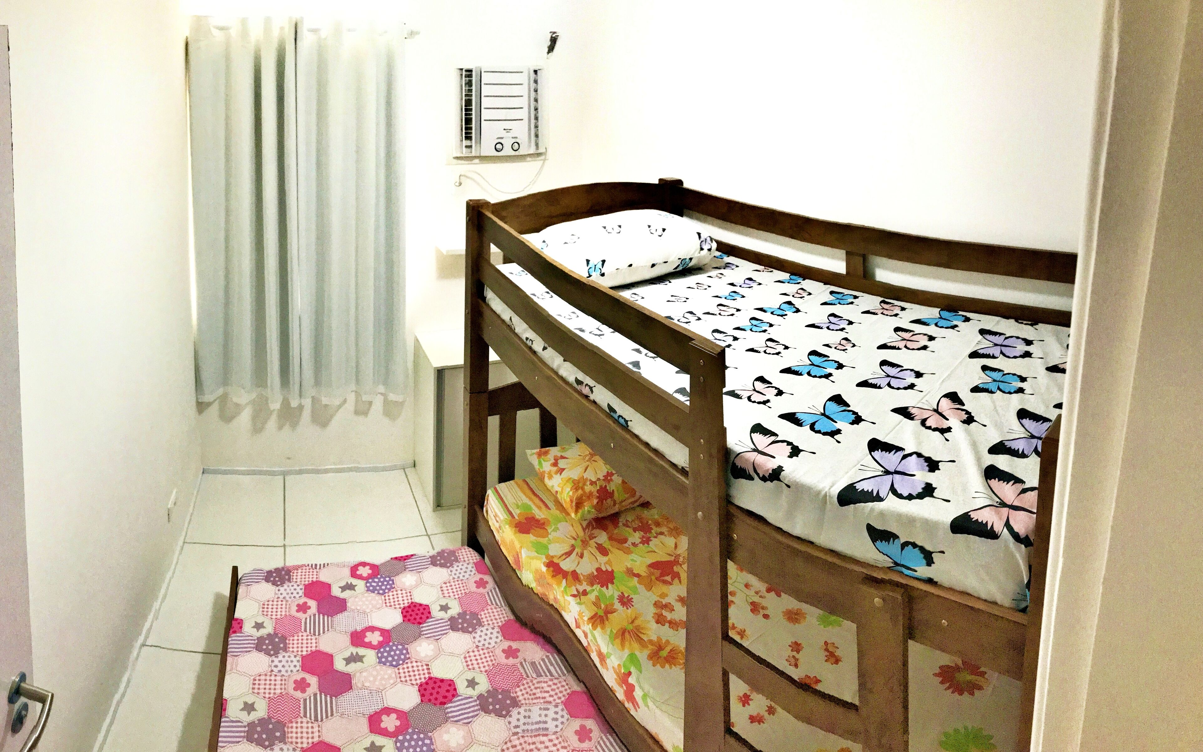 Piccola camera con un letto singolo a castello, una coperta a motivi floreali, un tappeto a pattern esagonale e una finestra con tenda.
