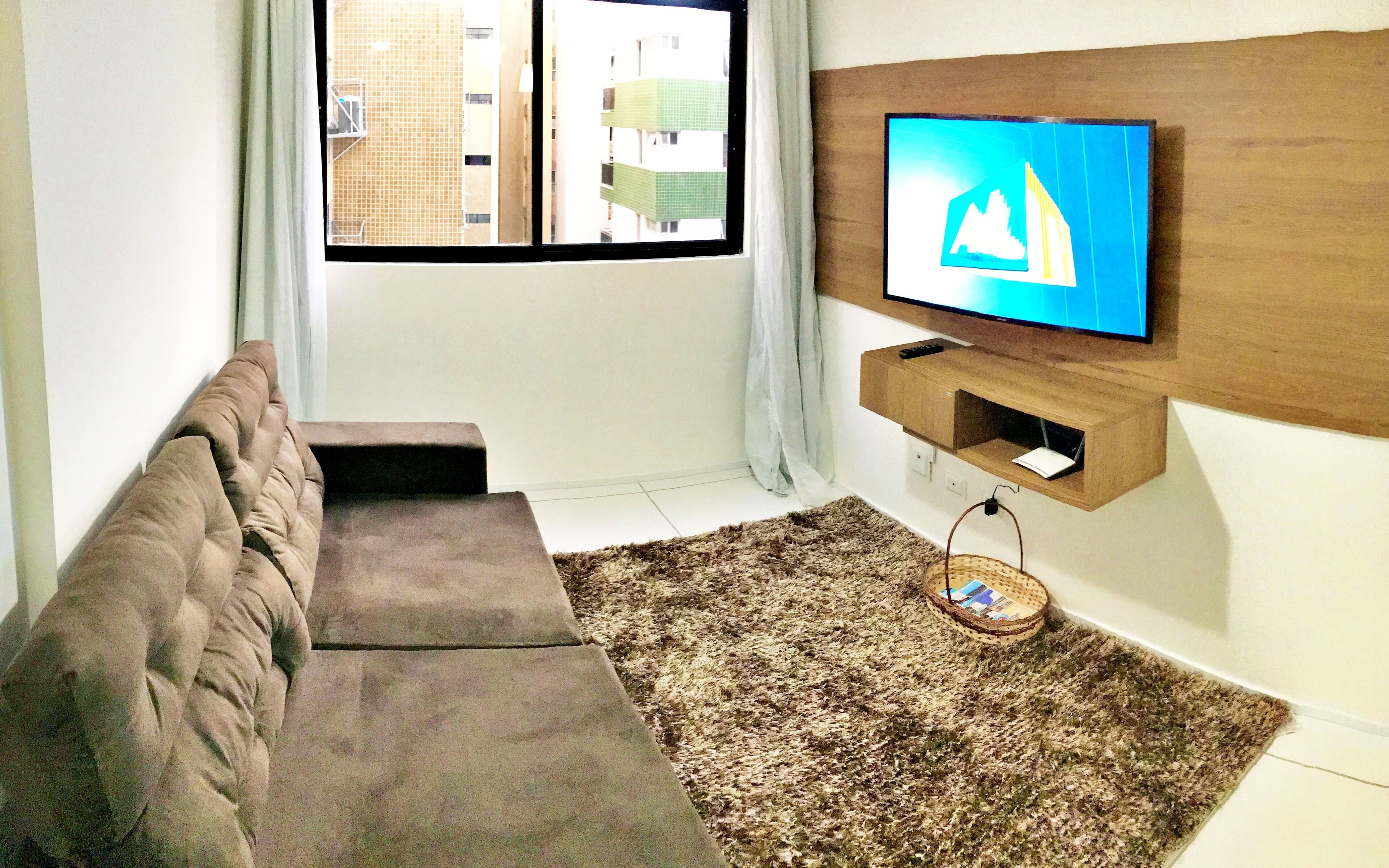 Un salon moderne avec un canapé, un téléviseur fixé sur un panneau en bois, une fenêtre donnant sur des immeubles et un tapis à motifs.
