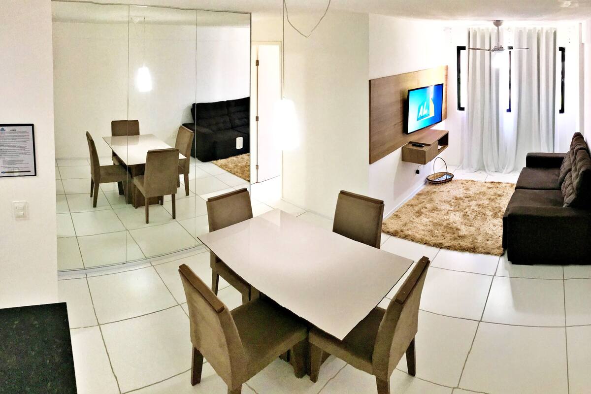 A modern living room with a dining area, a flat-screen TV, and a large window with curtains.