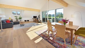 A spacious living area with a wooden floor, a sofa, a coffee table, and a dining table with chairs. There is a large window providing a view of the outdoors. - Landhotel Herzberger (Scheidegg)