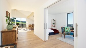 A spacious living area with a wooden floor, a sofa, a colorful armchair, and a bed with a red blanket. A large window offers a view of the outdoors. - Landhotel Herzberger (Scheidegg)