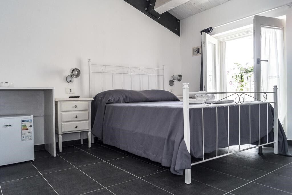 A bedroom with a white metal bed frame, a grey bedspread, a bedside table with a lamp, and a small white appliance.