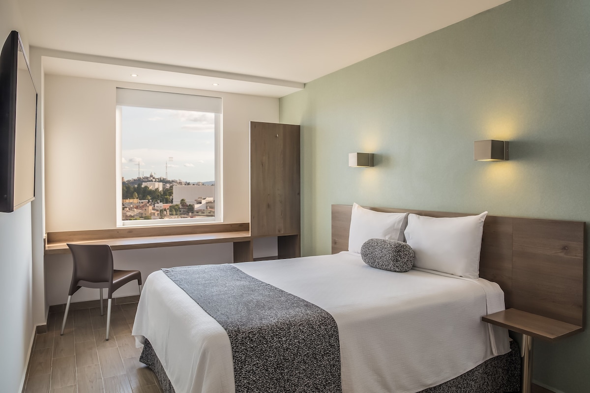 A modern hotel room with a large bed, a desk, and a chair. There is a window with a view of a cityscape.