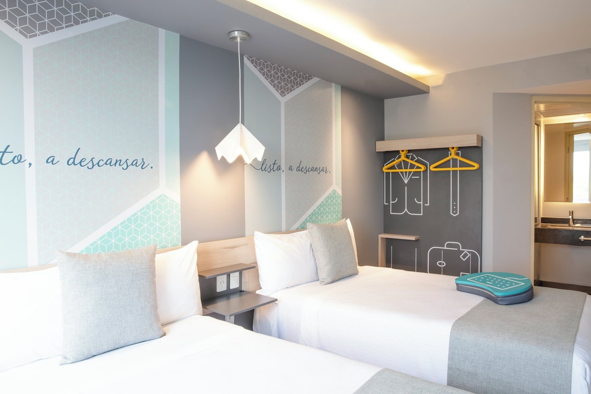 A hotel room with two beds, a desk, a hanging light fixture, a wall with geometric patterns, and a wardrobe with hangers.