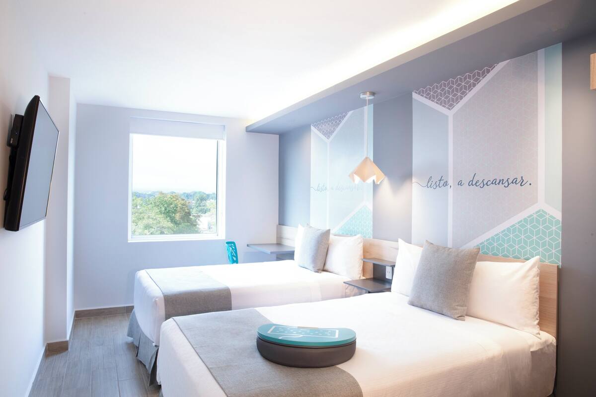 A modern hotel room with two beds, a large window, and a wall with geometric patterns and a motivational quote.