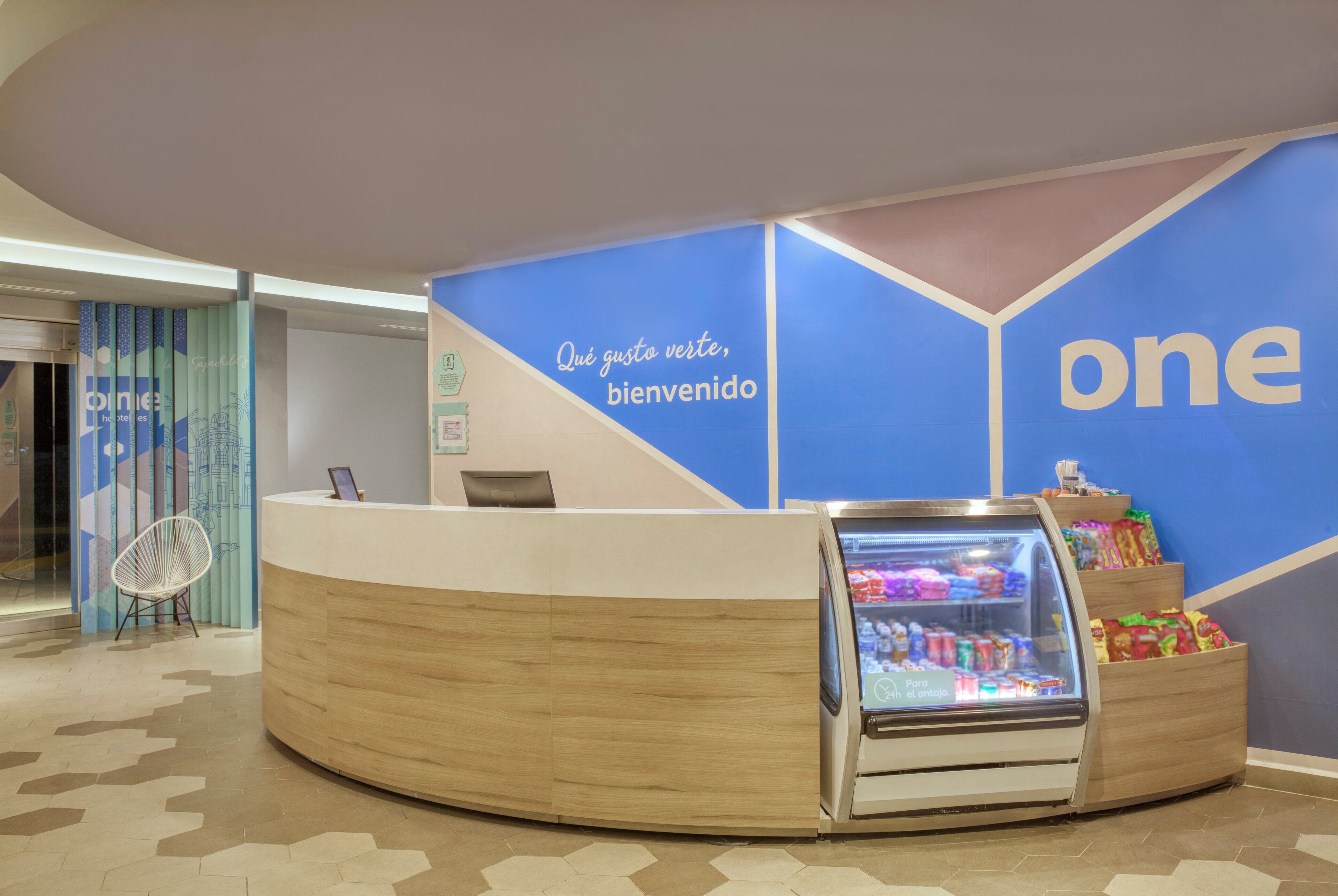 A reception desk with a digital screen, a refrigerator stocked with beverages, and a wall with the word 'one' and a welcome message.