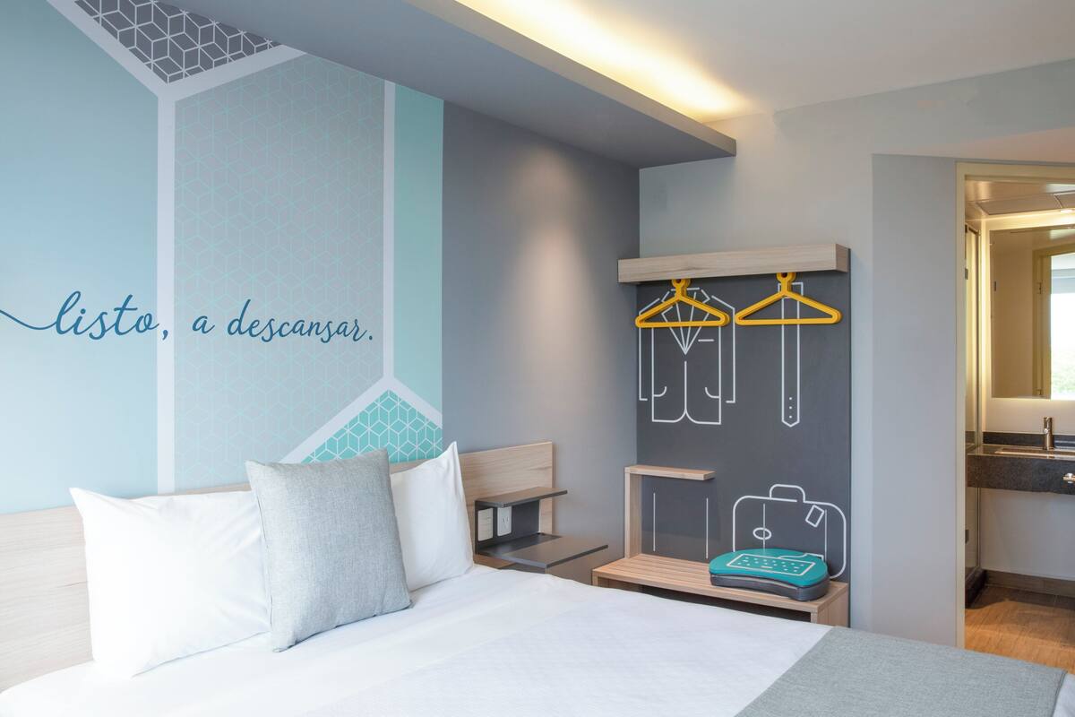 A modern hotel room with a bed, a wall-mounted coat rack, and a small table with a suitcase.