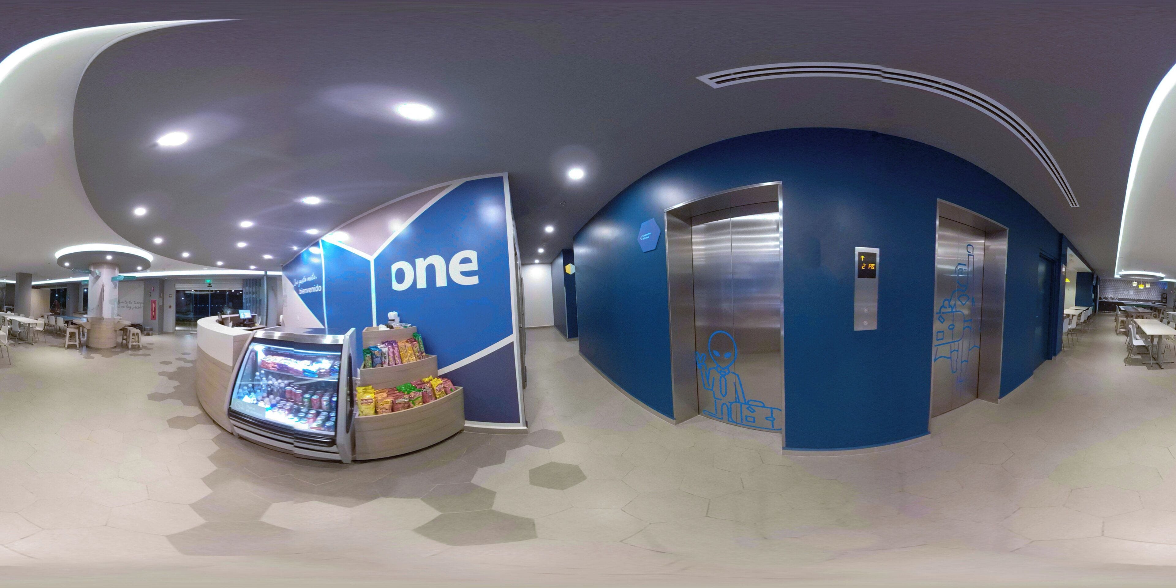 Un interior moderno con una paleta de colores azul y blanco, que incluye un quiosco con la marca 'one', un ascensor y una zona de estar con mesas y sillas.