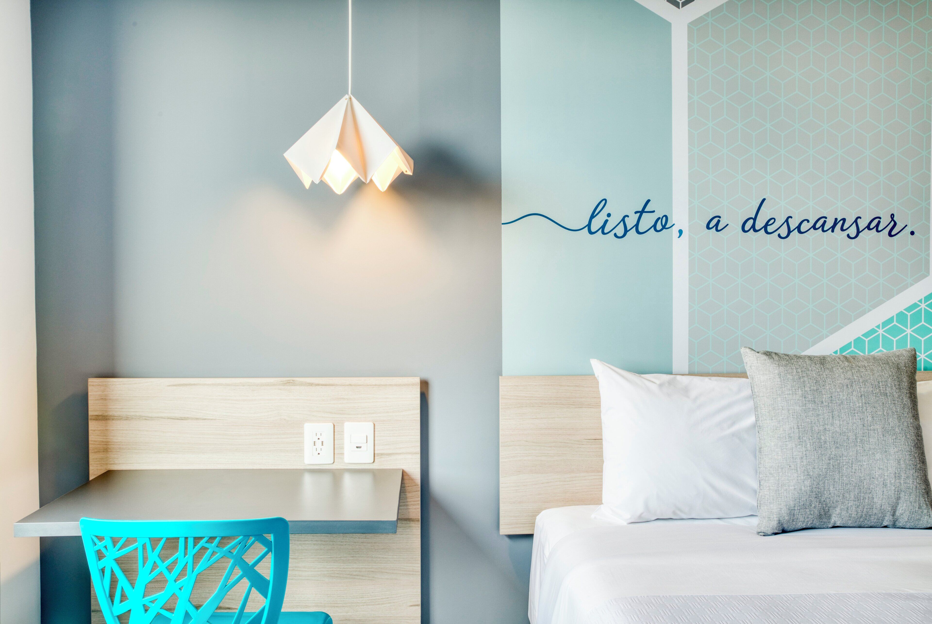 A modern room with a wooden headboard, a table, a chair, a bed with pillows, and a wall with a geometric pattern.
